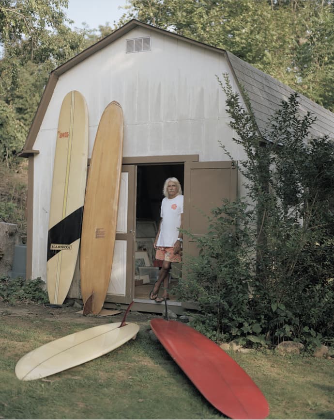 Photo from the book of a surfer by a barn with longboards and a red board