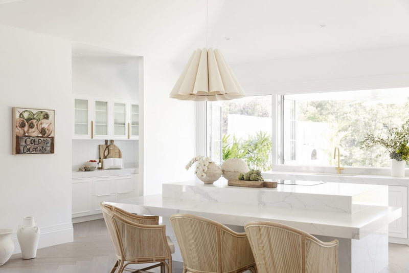 A white kitchen with wicker chairs and a white island, undergoing renovations with Three Birds Renovations: Dream Home How-To by Books to bring out its true potential.
