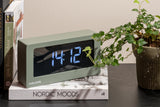 A Karlsson clock with a sleek design sits on a table next to a potted plant.