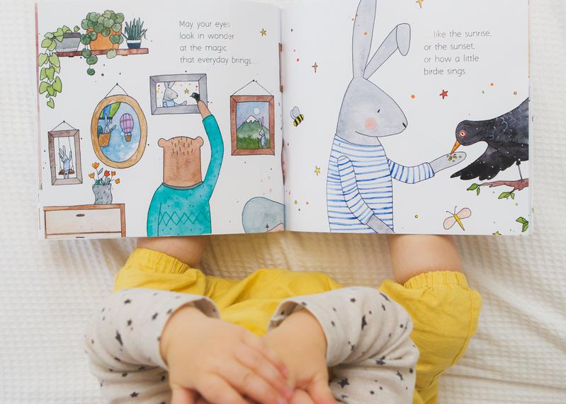 A baby is reading MY WISHES FOR YOU, a book by Olive + Page, with a picture of a bunny and a bird.