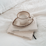 A cup of coffee sits on top of books on a bed, reflecting the Visions & Actions Journal and goals set in a personal journal by Collective Hub.