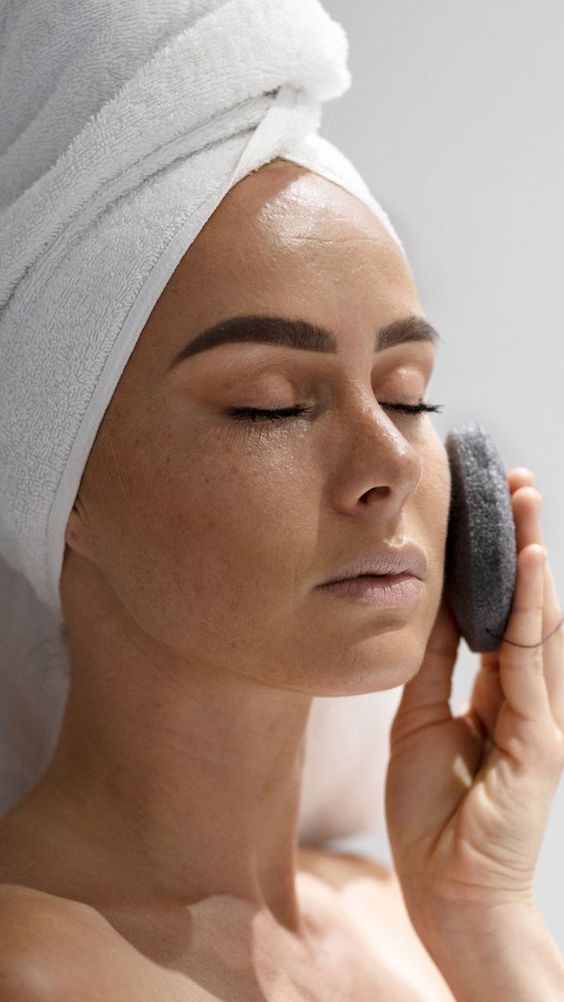 A woman in a towel is using a Florence Konjac Premium Facial Puff Sponge with Bamboo Charcoal to deep cleanse her face.
