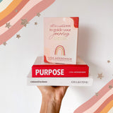 A woman holding up a "Affirmations to Guide Your Journey Box Card Set" by Collective Hub about purpose and affirmations.