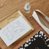 Limited edition Papier HQ stationery featuring an Abstract face notebook, pens, and a bottle of perfume on a wooden table.