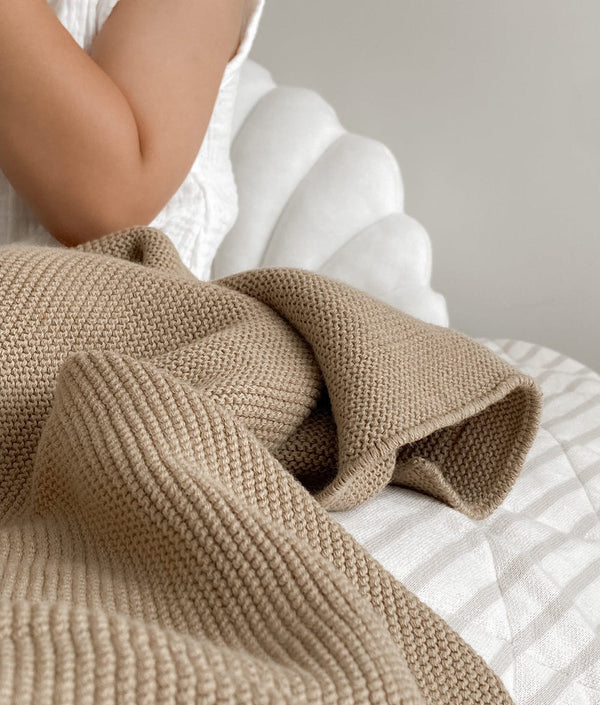 A woman is sitting on a bed with a Bengali Collections CLASSIC KNIT BLANKET - KHAKI.