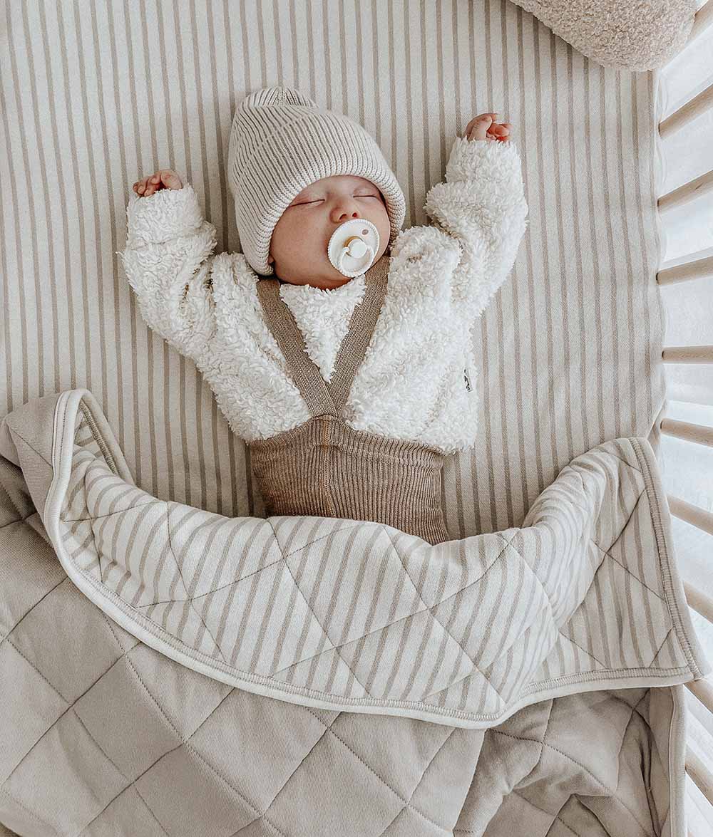 Baby asleep in a cot on a greige pinstripe jersey cot sheet under a quilted pinstripe blanket