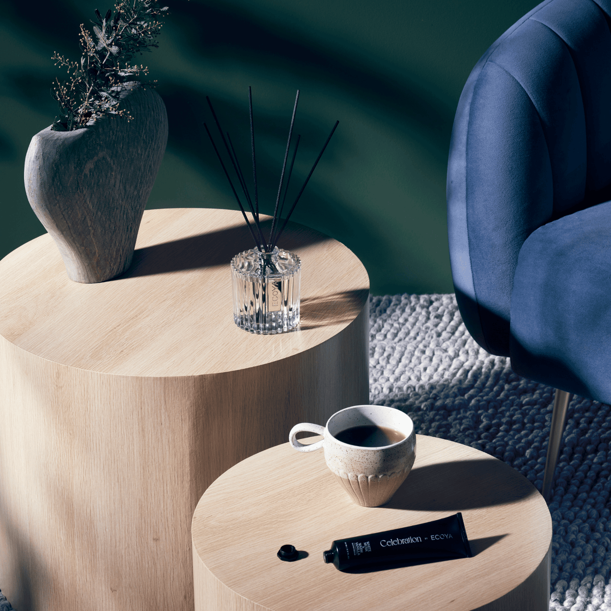 Celebration diffuser on a round timber side table beside a vase, with a coffee cup on a lower table