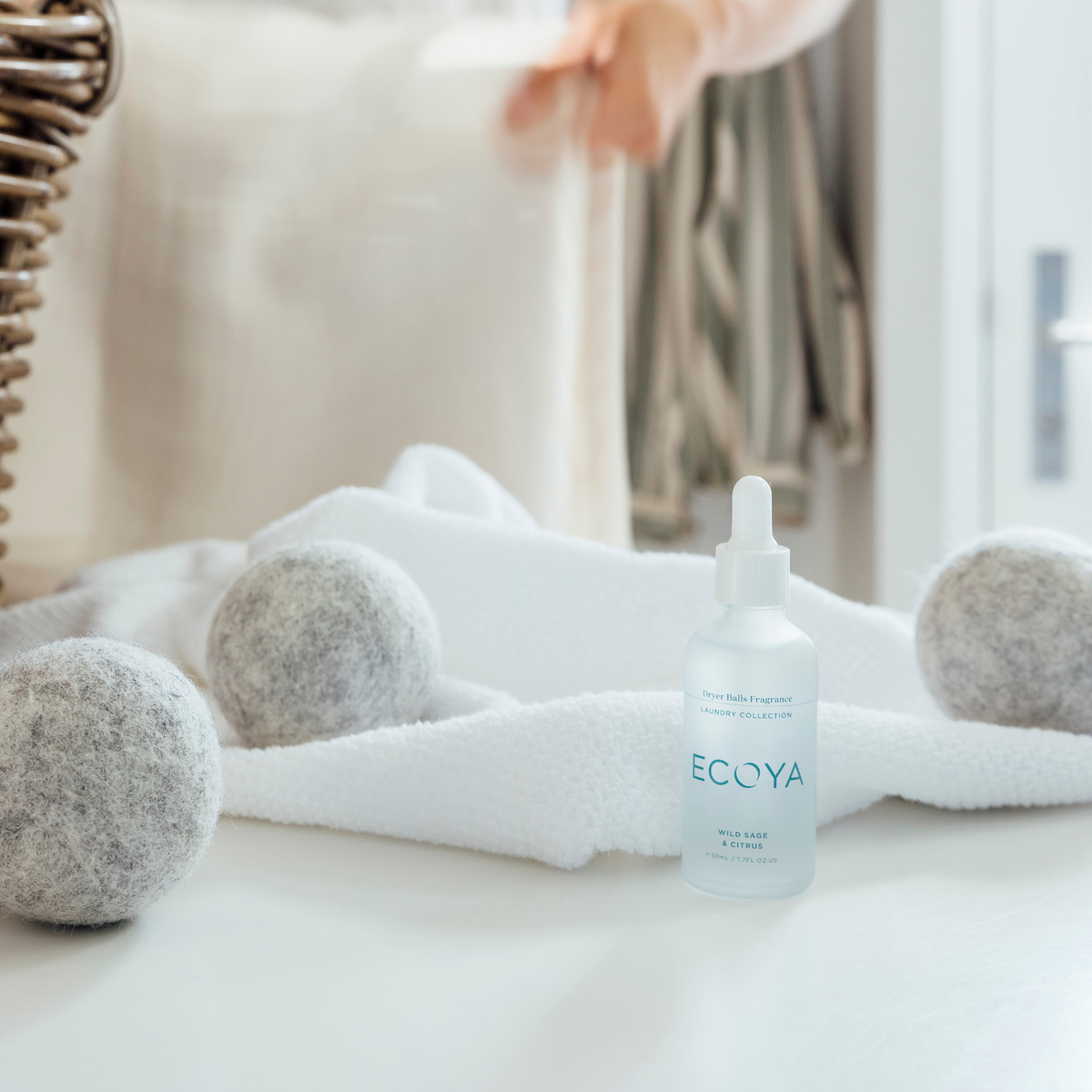 Folded white towels being placed in a basket with Ecoya wool dryer balls and fragrance dropper