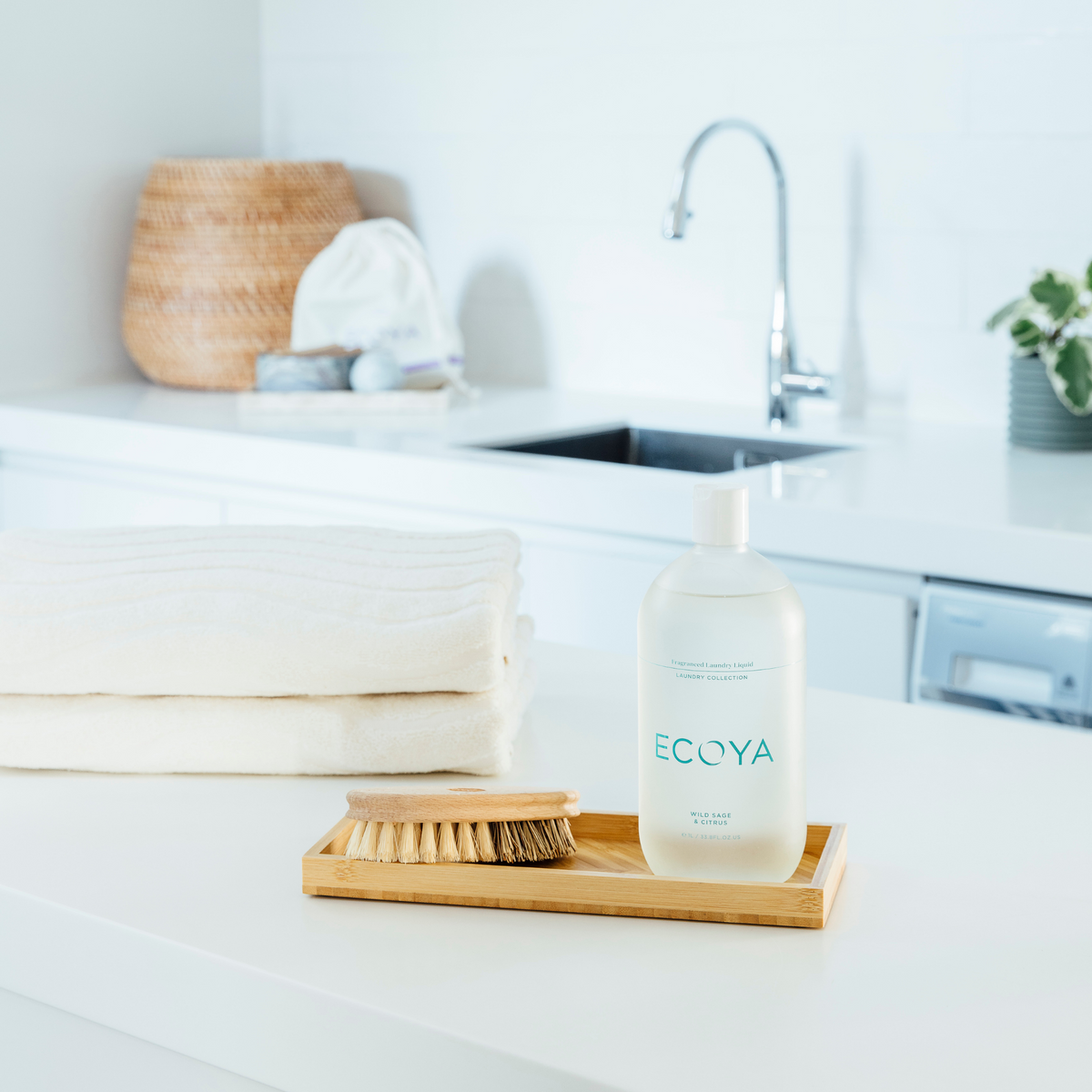 Folded white towels and Ecoya laundry liquid on a wooden tray beside a laundry sink