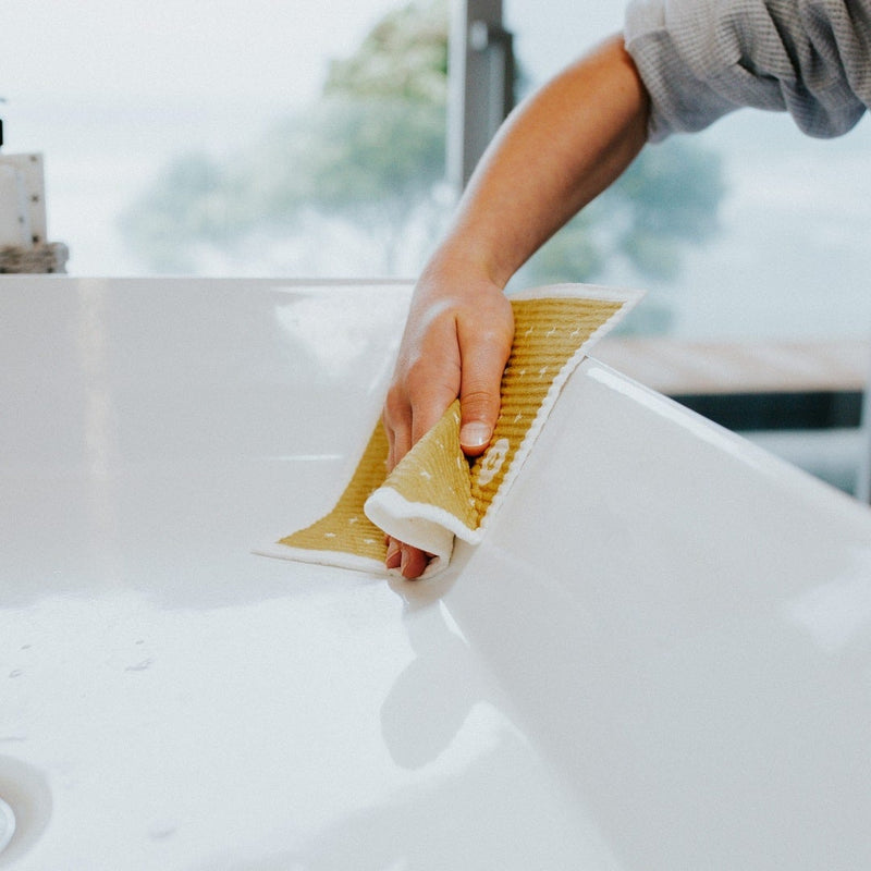 A person cleaning a sink with an ECO CLOTH- MEDIUM (3-PACK) sponge, using Good Change modern designs.