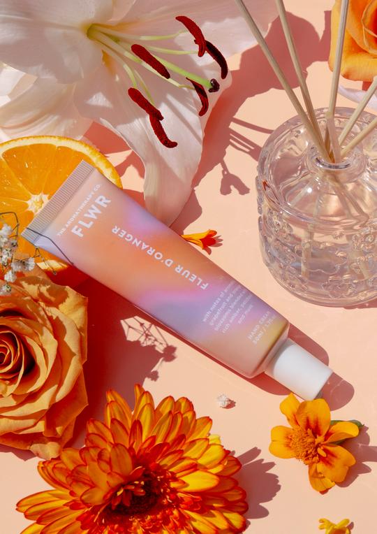 A tube of FLWR Hand Cream - Fleur D'Oranger by The Aromatherapy Co and orange flowers on a pink background.