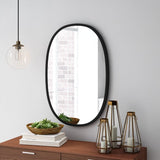 The Umbra Hub Mirror Oval - Black features a sleek black design, suspended above a wooden dresser, with a stylish rubber rim for added elegance.