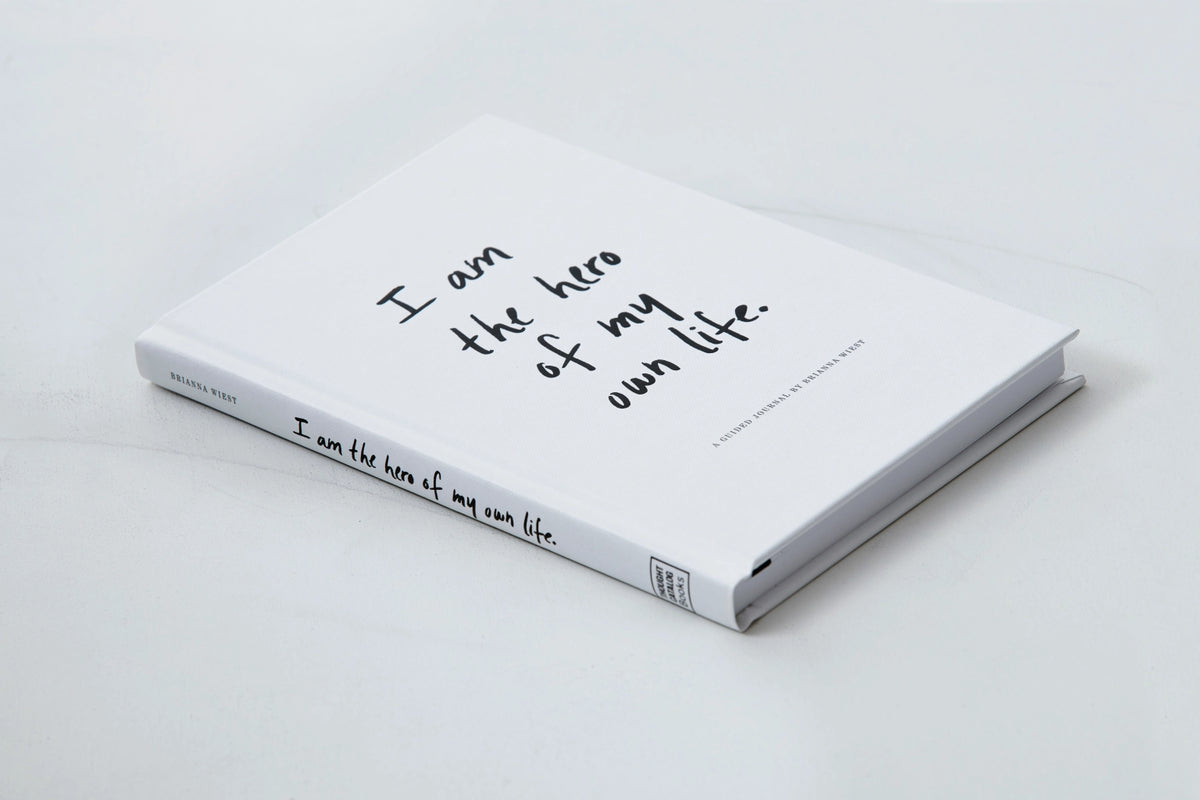 A book on design and lifestyle, "I Am The Hero Of My Own Life" by Brianna Wiest and published by Thought Catalog, that helps you envision your ideal life and overcome unconscious attachments with the words "I am the light of my life" written on it.