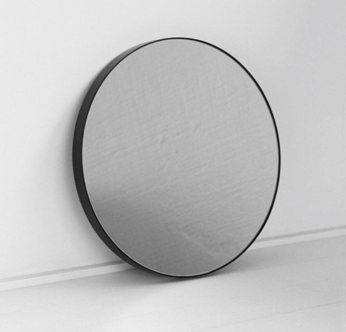 Round black frame mirror leaning against a white wall on a pale floor