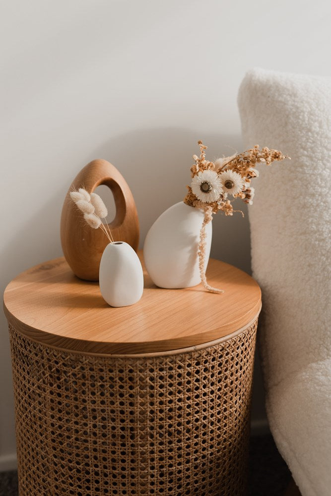 The Great Harmie Vase - White / Natural, handmade by Vietnamese artisans, adorns a wicker side table in the Ned Collections.