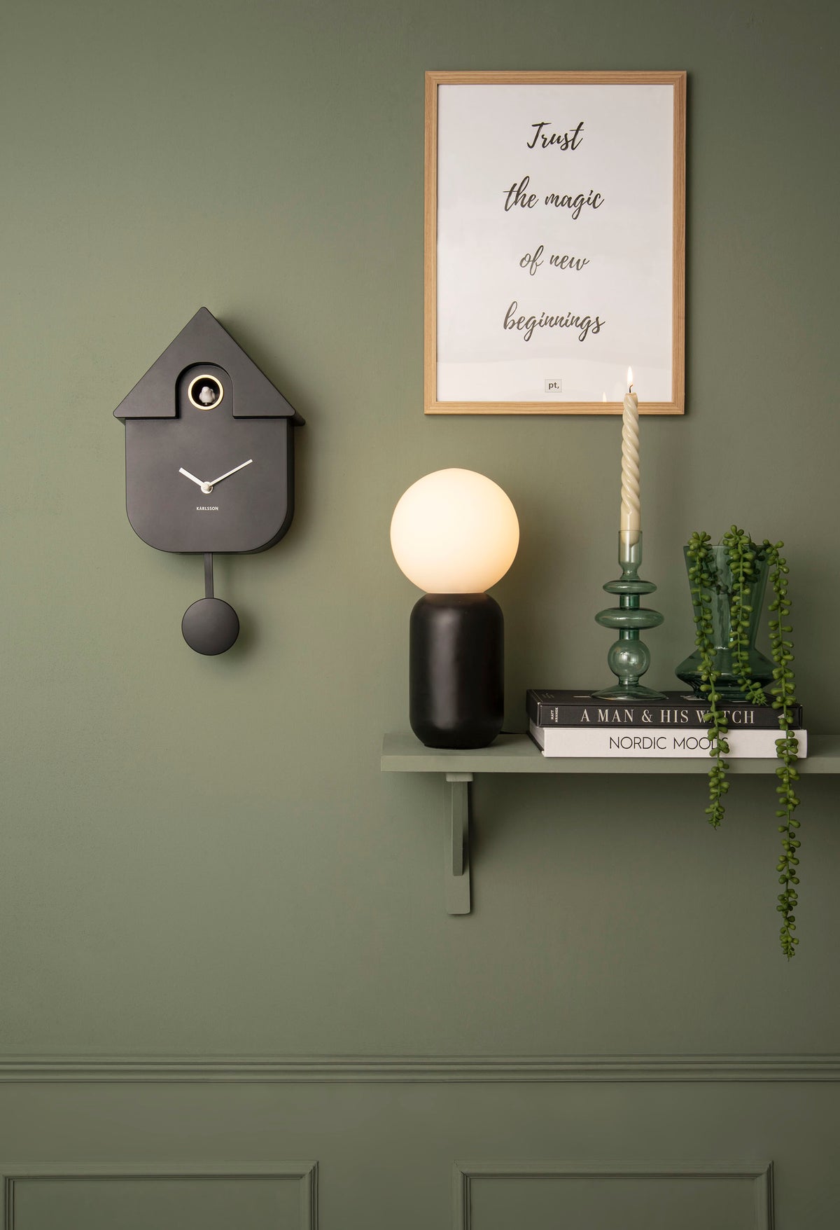 Karlsson Modern Cuckoo Clock in black resting on a minimal shelf against a green wall