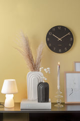 A Karlsson Charm Wall Clock - Various Colours featuring engraved gold numbers, displayed on a yellow wall.