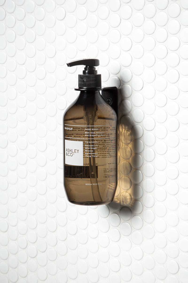A bottle of soap, specifically the Made of Tomorrow FOLD Bottle Holder ∙ Black, sitting on a white tile wall in a bathroom or office setting.