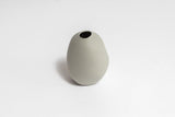 A small grey Harmie Vase - Seed Grey, handmade by Vietnamese artisans, sitting on a white surface. 
Brand: Ned Collections
