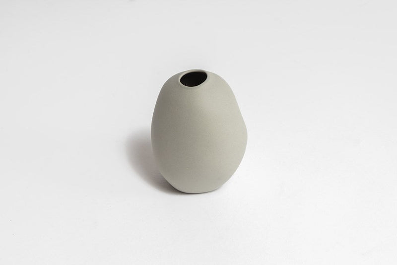 A small grey Harmie Vase - Seed Grey, handmade by Vietnamese artisans, sitting on a white surface. 
Brand: Ned Collections