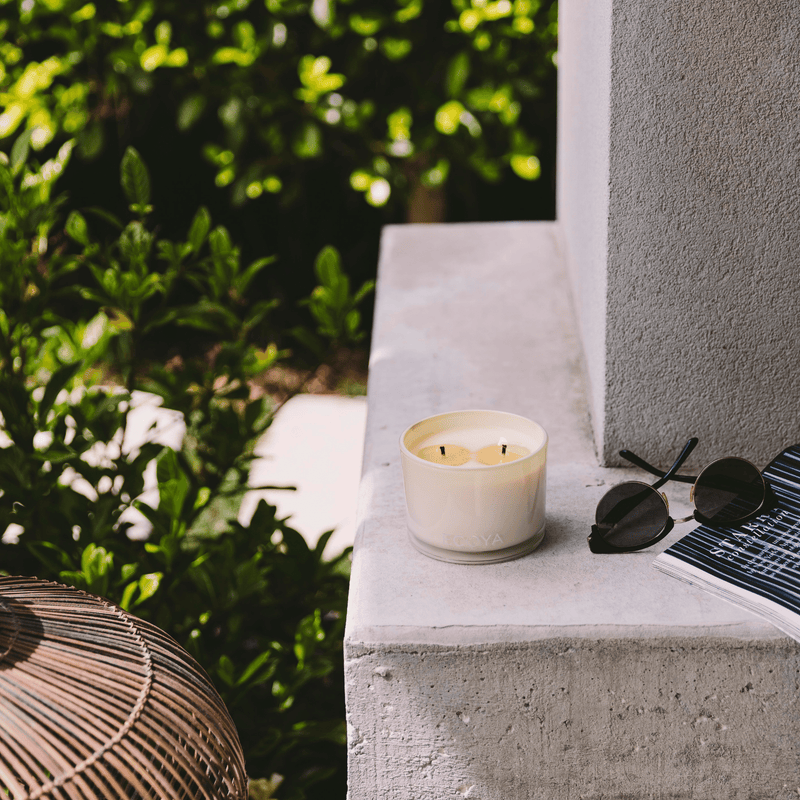 A Scandinavian-inspired Ecoya Limited Edition Citronella & Lemongrass Outdoor Candle adds a touch of home design and home fragrance to the serene ambiance, alongside a book and sunglasses on a ledge.