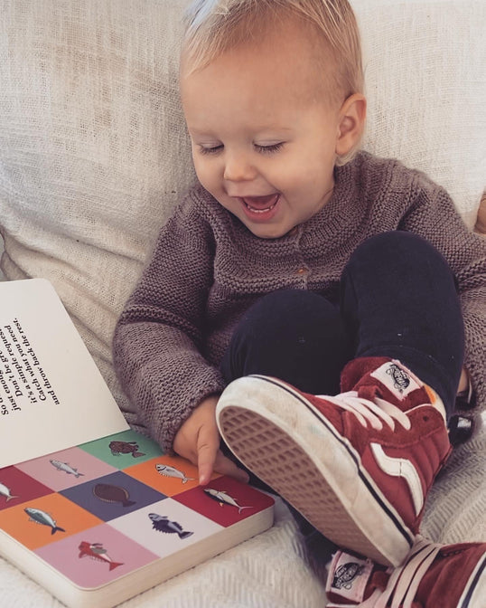 A baby is sitting on a couch reading the Fish of Aotearoa book by As We Are Illustration.
