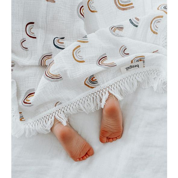 A baby's feet are laying on a bed wrapped in a white Muslin Fringe Swaddle - Rainbow blanket made from Oeko-tex® Certified Cotton, by Bengali Collections.