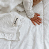 A baby laying on a white Bengali Collections White Heirloom Blanket with a teddy bear.