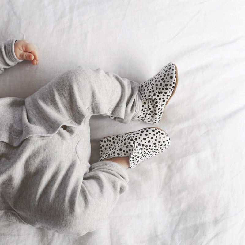 A baby is laying on a bed wearing LTL BIG | URBAN SLIP-ON IN MESSY SPOT shoes made of natural leather.