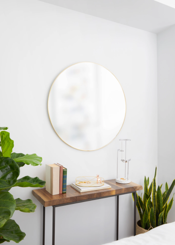 A white bedroom with a round Umbra HUBBA MIRROR 86cm BRASS wall mirror and a plant on the table.