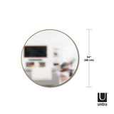 A HUBBA MIRROR 86cm BRASS by Umbra, a decorative wall mirror with a metal frame, is shown in front of a living room.