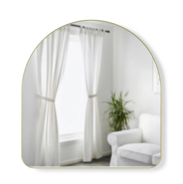 A HUBBA ARCHED MIRROR - BRASS by Umbra with a metallic finish rim and unique arch shape adds a decorative touch to a room with white curtains and a white couch.
