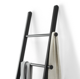 The Umbra LEANA LADDER - BLACK provides a stylish storage solution, featuring a sleek black design and a convenient towel hanging on it. Perfect for optimizing space and adding a touch of elegance to any room.