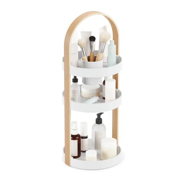 An Umbra BELLWOOD COSMETIC ORGANIZER with a three-tier bathroom shelf filled with cosmetics and makeup brushes.