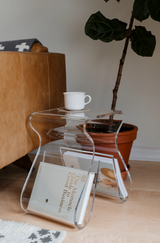 A Magino Stool with Magazine Rack - Clear Acrylic by Umbra with books and a plant on it.
