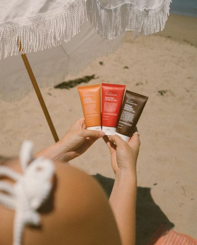 A woman holding three tubes of We Are Feel Good Inc. Coconut Sunscreen SPF 50+ lotion on a beach.