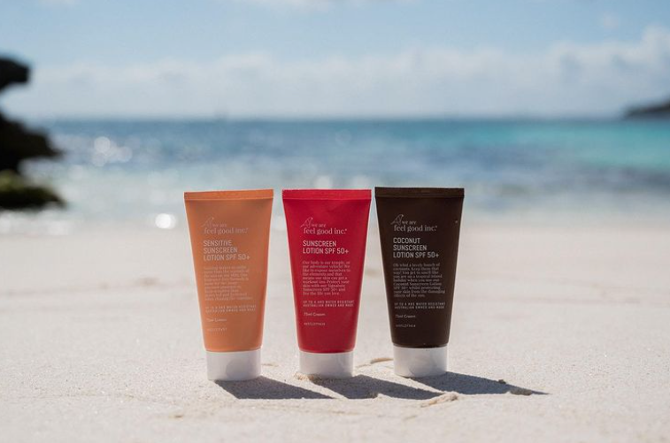 Three tubes of We Are Feel Good Inc. Coconut Sunscreen SPF 50+ sitting on a sandy beach.