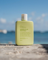 A bottle of We Are Feel Good Inc. Kakadu Plum Body Milk sitting on a wooden table in front of the ocean.
