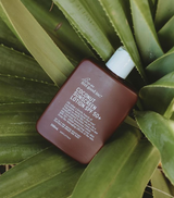 A bottle of We Are Feel Good Inc. Coconut Sunscreen SPF 50+ sitting on top of a plant.