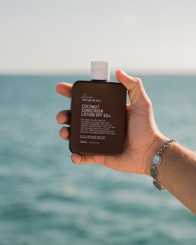 A hand holding a bottle of We Are Feel Good Inc. Coconut Sunscreen SPF 50+ in front of the ocean.