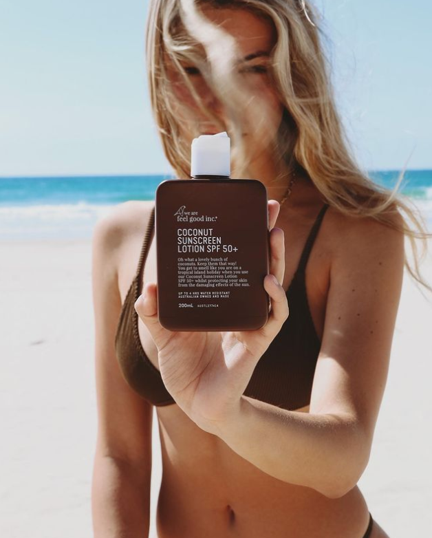 A woman in a bikini holding a bottle of We Are Feel Good Inc. Coconut Sunscreen SPF 50+ on the beach, ensuring reef-safe protection with broad spectrum.