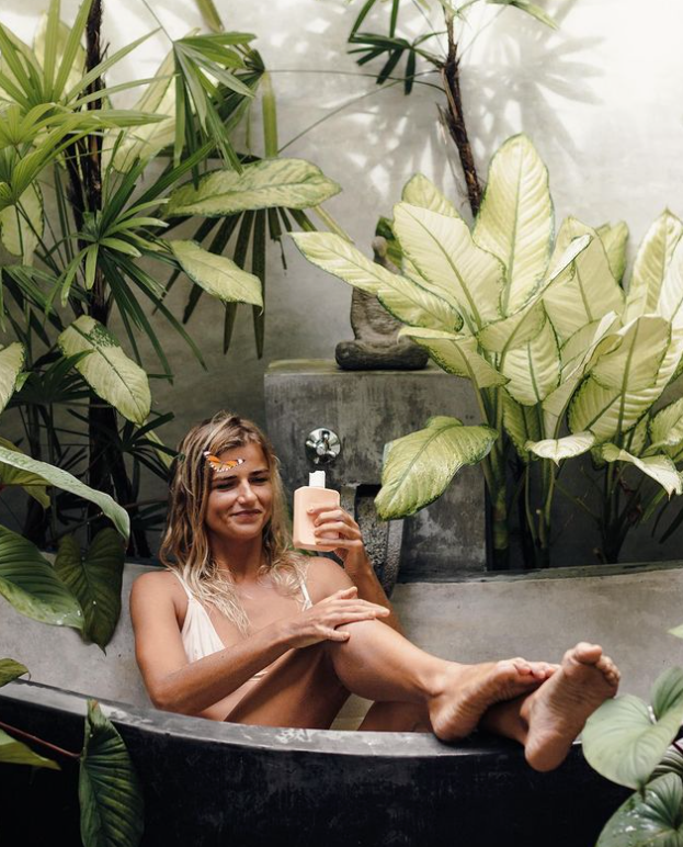 A woman sitting in a tub, surrounded by plants, while using We Are Feel Good Inc.'s Sensitive Body Milk for sensitive skin.