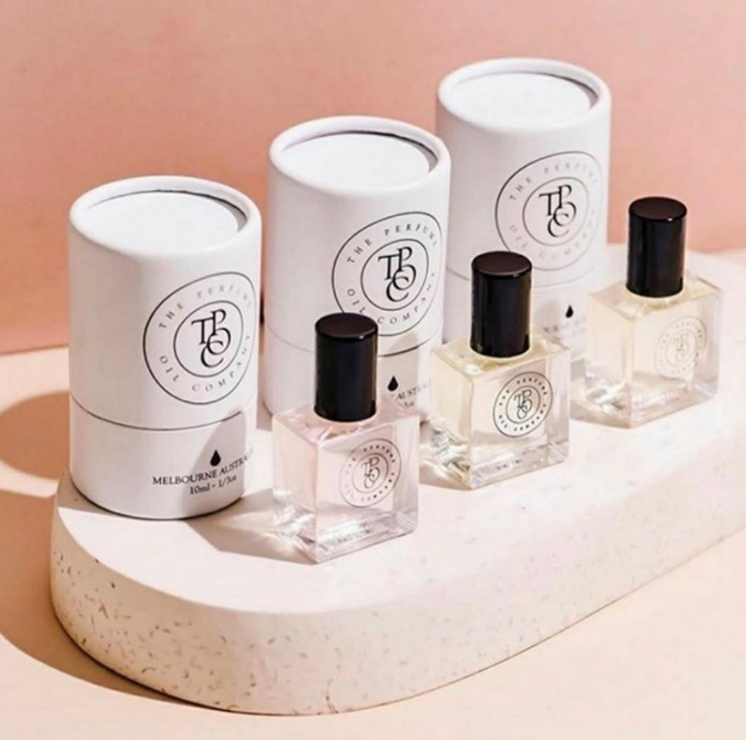 Three perfume oil bottles and gift tubes on a white plinth against a blush pink backdrop