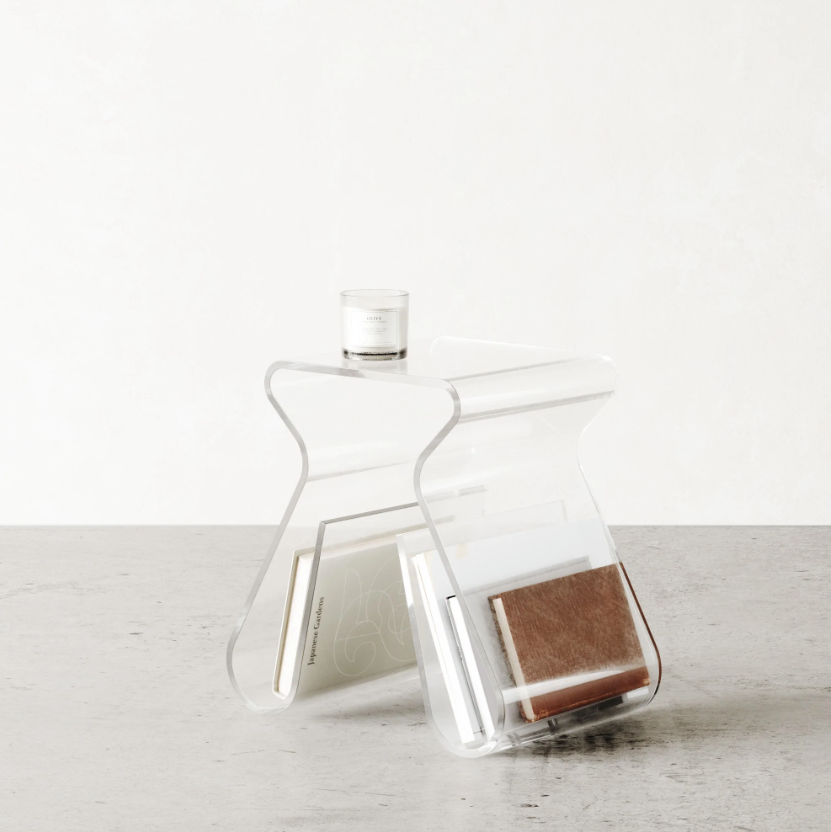 Clear acrylic Magino stool holding a book in its rack, a glass on top, against a pale wall