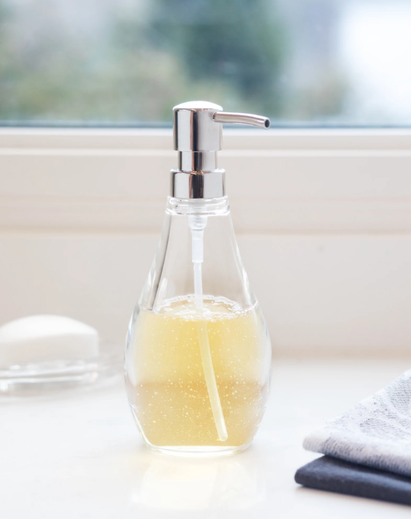 A Droplet soap pump, clear, featuring the Umbra design, sitting on a window sill.
