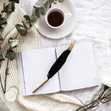 An open AXEL & ASH notebook adorned with a Black Feather Pen, accompanied by a steaming cup of coffee for a cozy writing experience.
