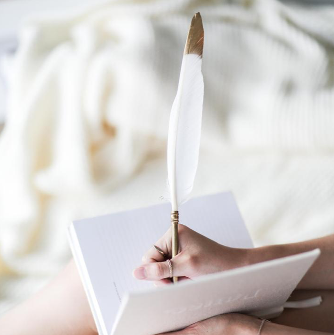 Hand writing in a white notebook with the Coco feather pen against soft cream fabric
