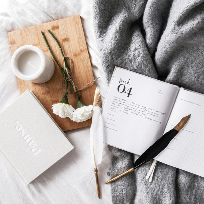 Coco feather pens with a notebook, coffee and white flowers on a timber board and grey throw