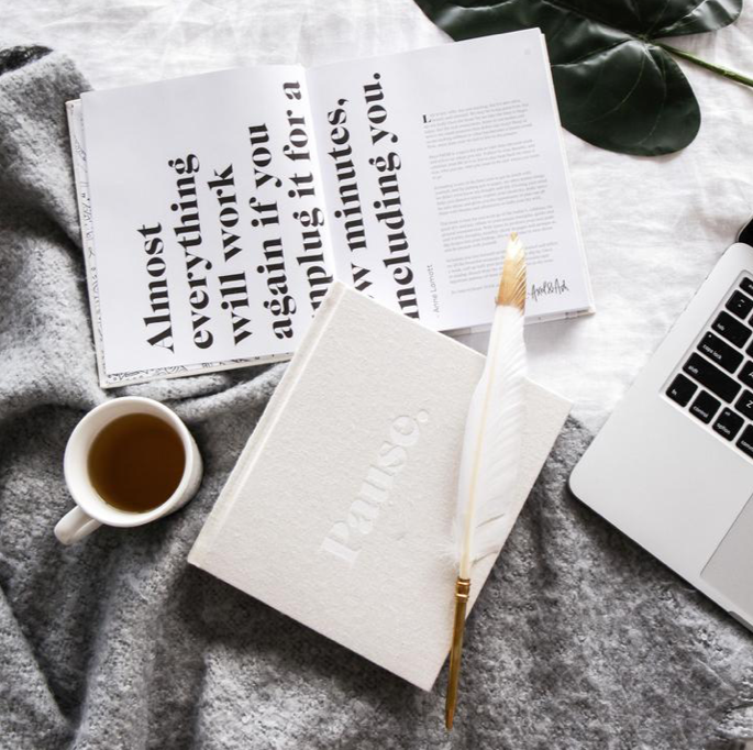 Coco feather pen on a notebook with coffee, a laptop and an open magazine on a grey throw