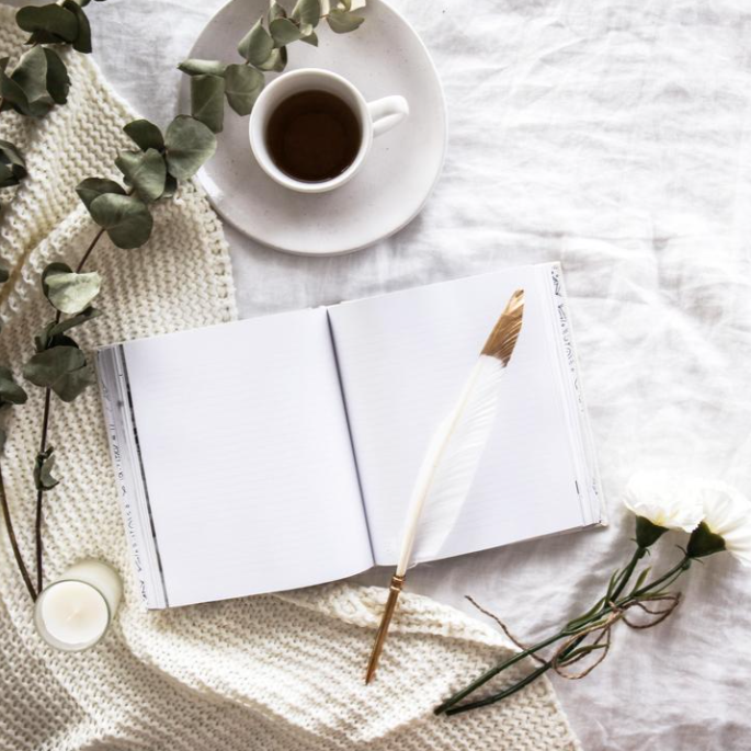 Coco feather pen resting on an open notebook beside coffee, eucalyptus and a knit throw